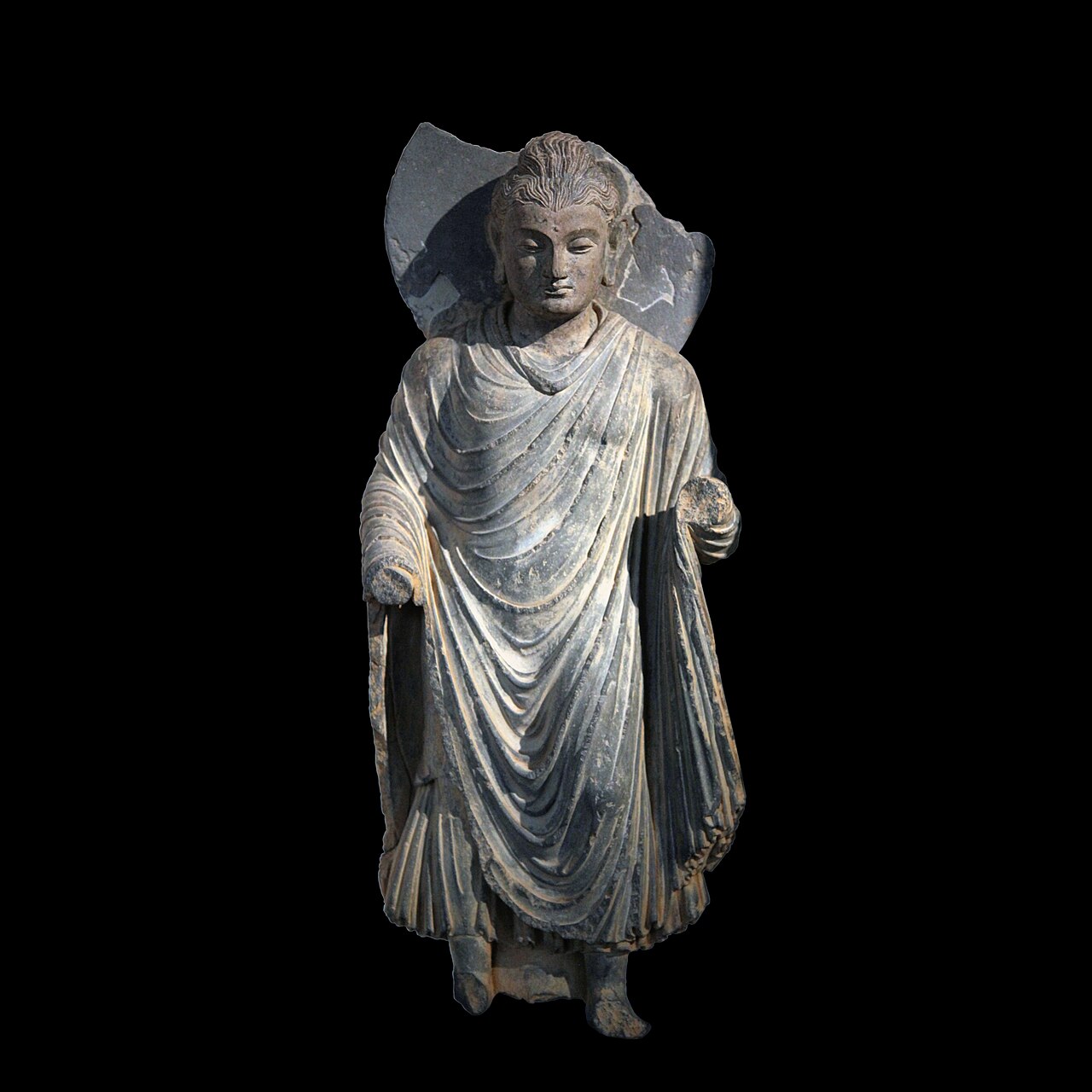 A standing Gandhara Buddha in grey schist, wearing heavy classical drapery with deep folds, photographed against a black background.
