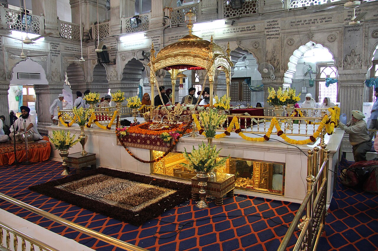 Inside Gurdwara Sis Ganj Sahib in Delhi, the Guru Granth Sahib rests draped and garlanded under a gilded canopy on its throne while an attendant waves the chaur whisk over it.