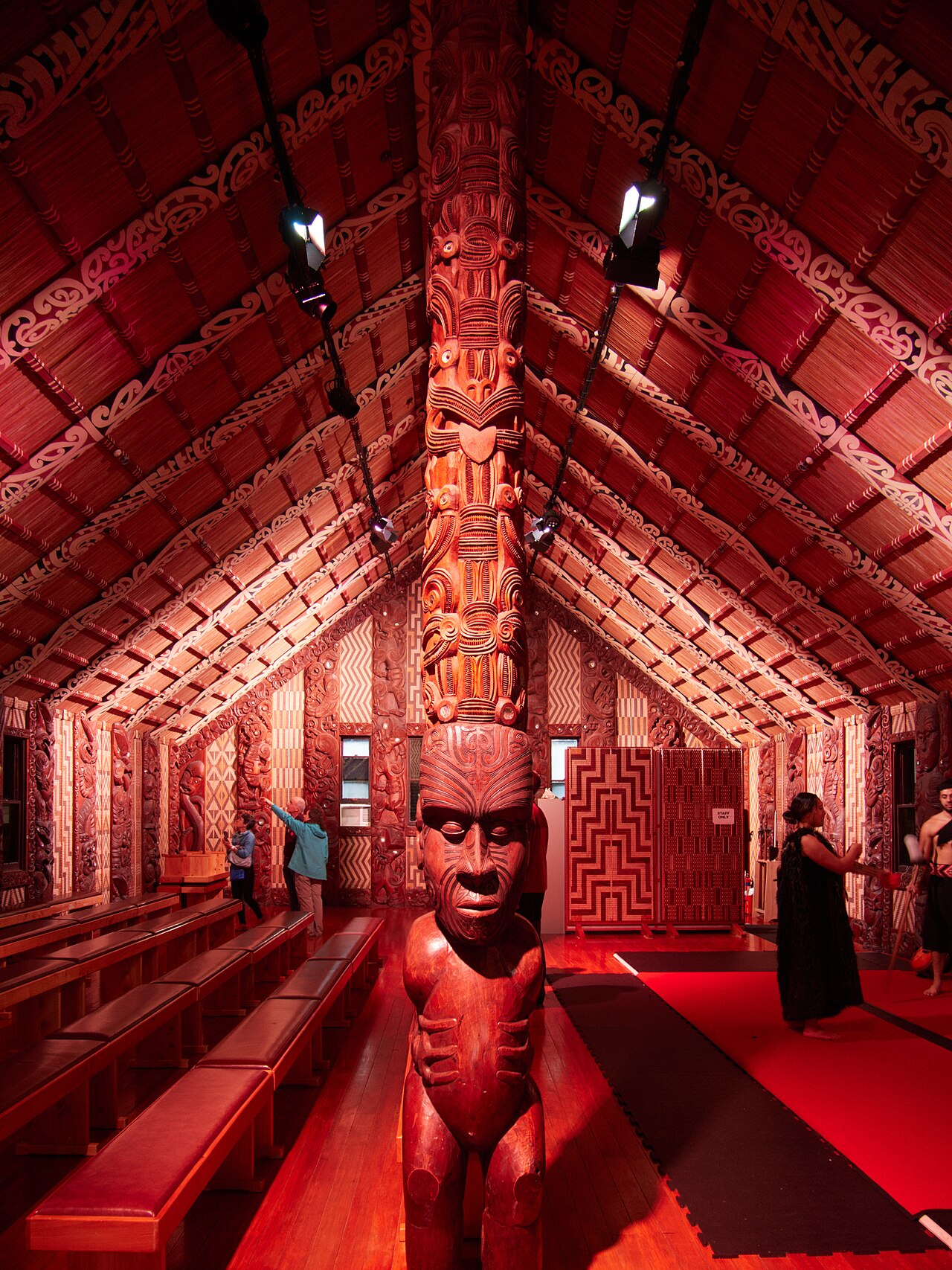 Inside the wharenui at Waitangi: red-carved rafters, woven tukutuku panels, and the tekoteko post of the meeting house at the Treaty Grounds.