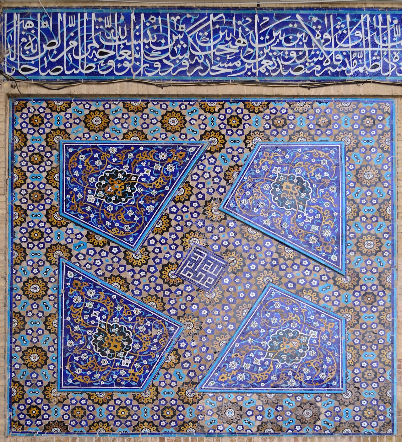 A mosque wall panel in Isfahan covered with blue geometric star tiles and floral arabesque, topped by a band of white thuluth Arabic calligraphy on deep blue.