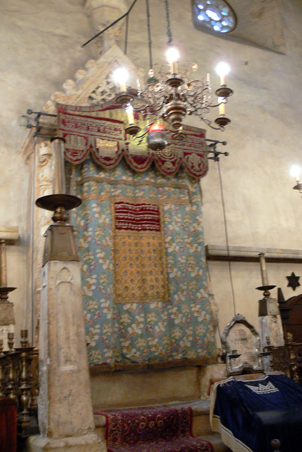 The Torah ark of Prague's Old-New Synagogue, its stone gable hung with an embroidered curtain (parochet) beneath a hanging lamp and chandelier.