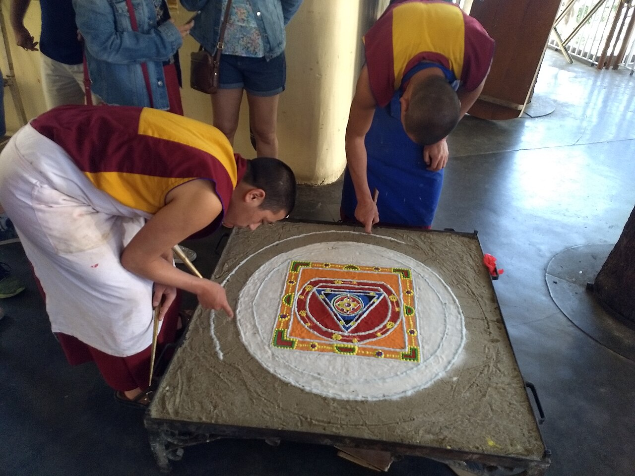 Two Tibetan monks bent over a table, laying colored sand through metal funnels to build a circular mandala with a square palace plan at its center.