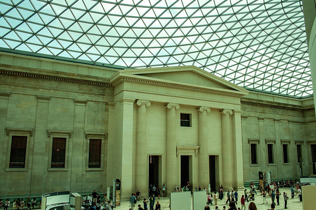 Visitors crossing the British Museum's Great Court beneath its tessellated glass roof, facing the classical stone façade of the Reading Room.