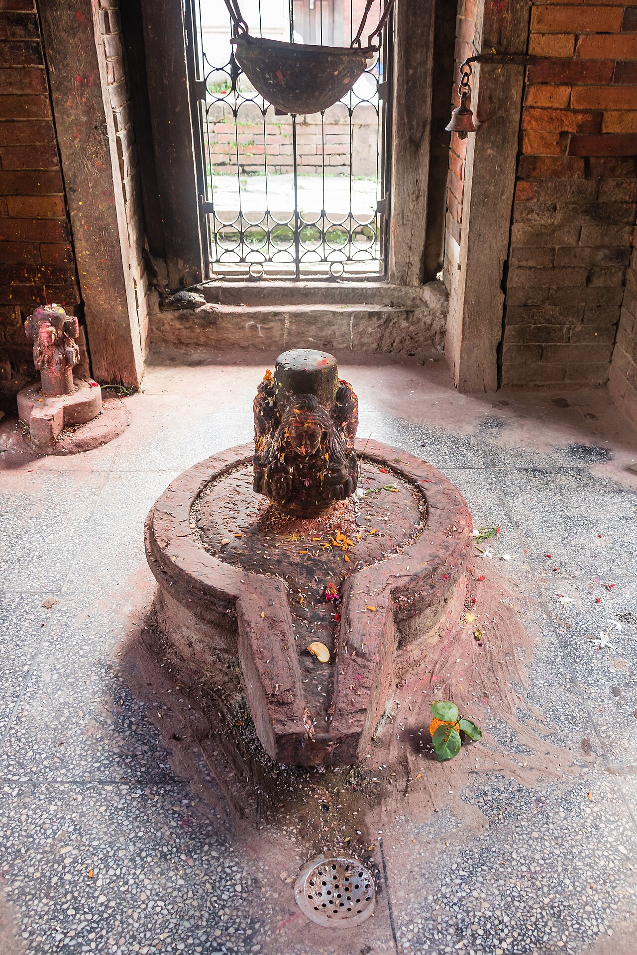 A stone Shiva linga on its round yoni base inside a small temple shrine, scattered with flower petals and pigment, a drip vessel hanging above.