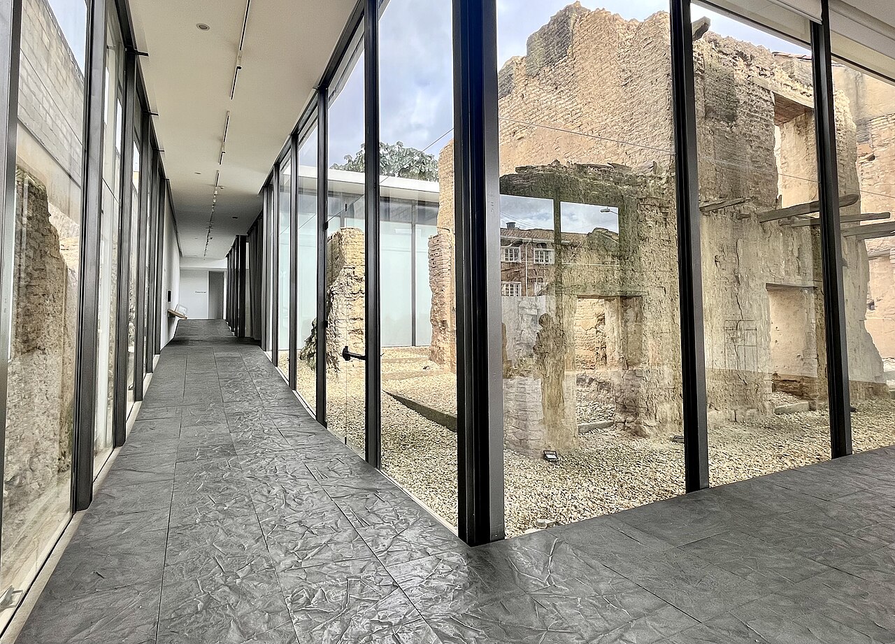 Inside Doris Salcedo's Fragmentos memorial in Bogotá: a glass-walled corridor whose floor is made of her textured metal plates cast from melted-down weapons, with preserved ruins visible through the glass.
