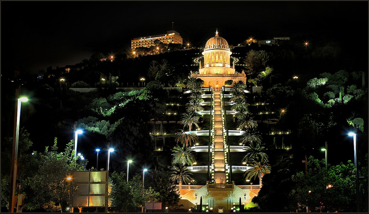 A night photograph of the Bahá'í Terraces on Mount Carmel in Haifa: the illuminated golden-domed Shrine of the Báb at the top and the lit central staircase and terraces descending between palm trees.