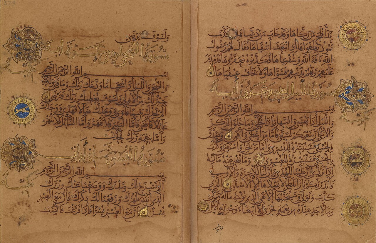 An open two-page spread of the Qur'an copied by Ibn al-Bawwab in 1000-1001, written in flowing cursive script with gold chapter headings and verse medallions.