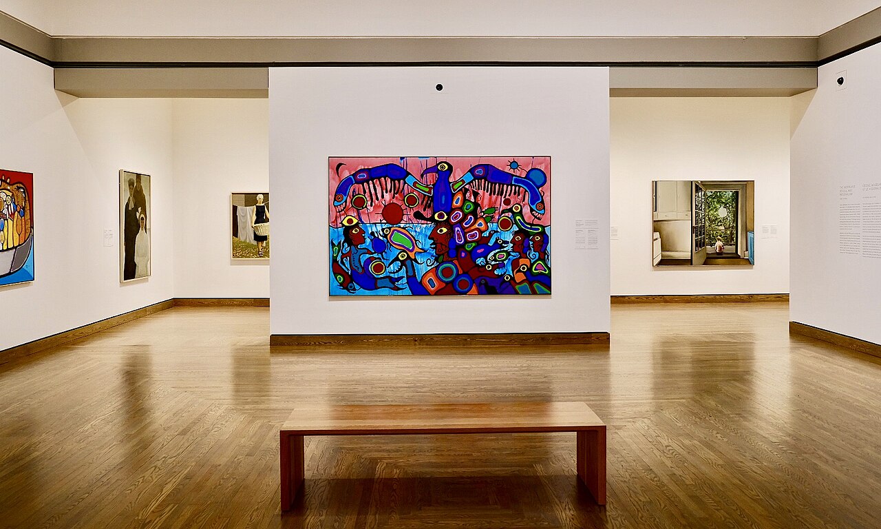 A gallery interior photograph showing Norval Morrisseau's large painting Artist and Shaman Between Two Worlds — a blue thunderbird spreading its wings over a dense field of figures and spirit beings in red, pink, and turquoise — hanging on a museum wall above a viewing bench.
