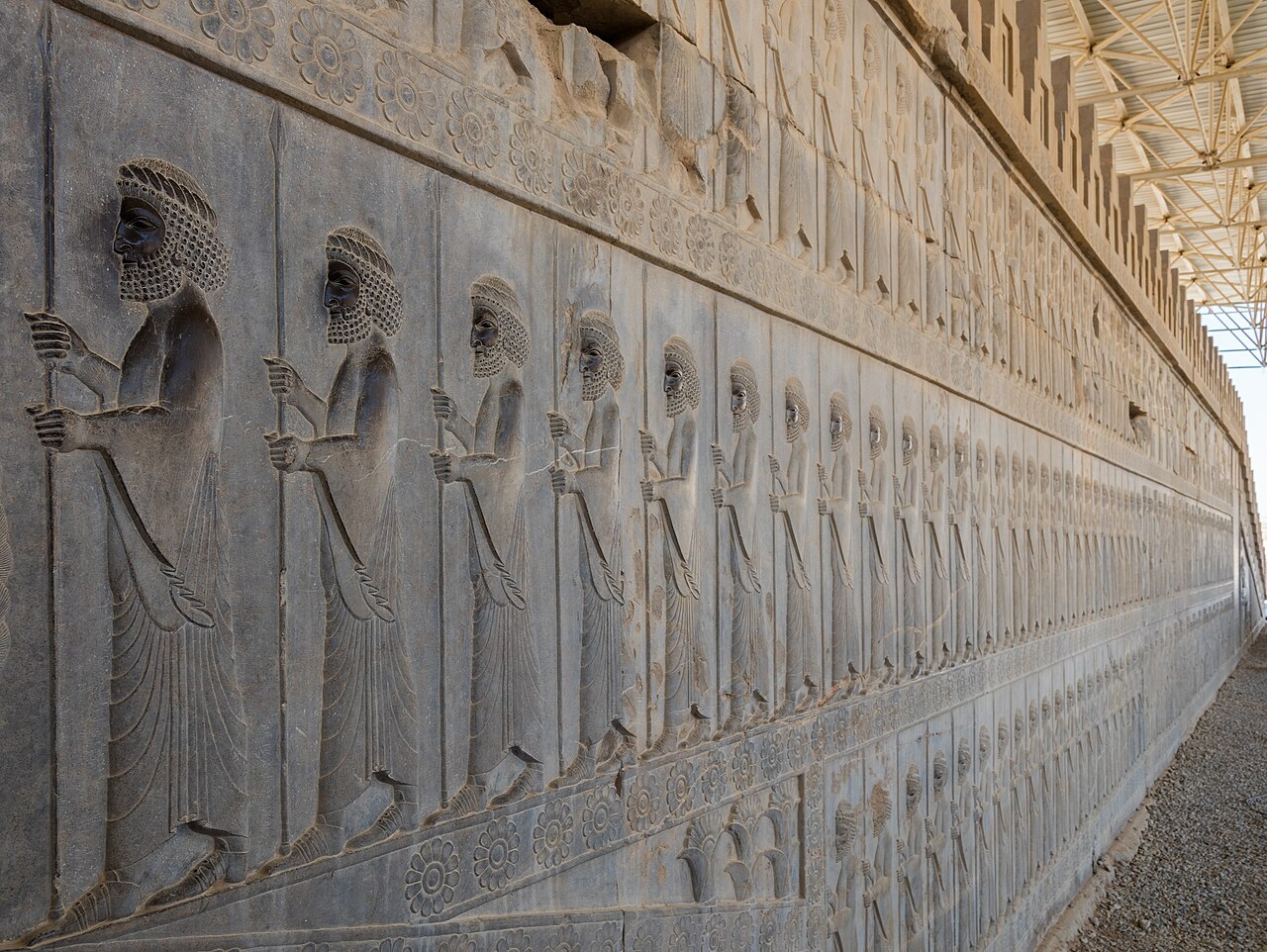 A photograph along the Apadana relief wall at Persepolis: a long receding procession of carved Persian guards and tribute bearers in profile, with rosette friezes above and below.