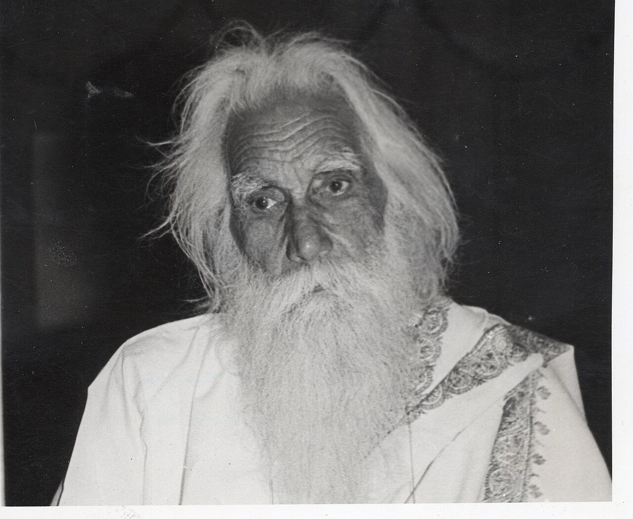 A 1985 black-and-white portrait photograph of Sobha Singh in old age, his long white hair loose around his shoulders and a long white beard over a white robe with an embroidered shawl edge.