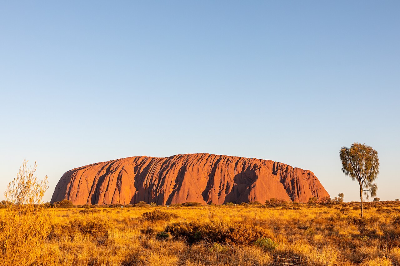 Uluru glows red-orange in low sunlight above flat desert scrub, seen from a public viewpoint.