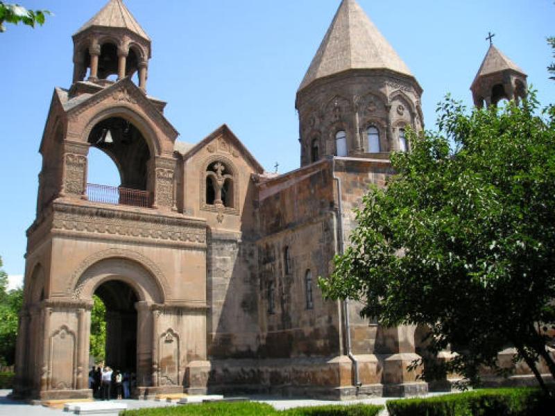 Etchmiadzin Cathedral's stone facade, conical dome, and open belfry under a blue sky.