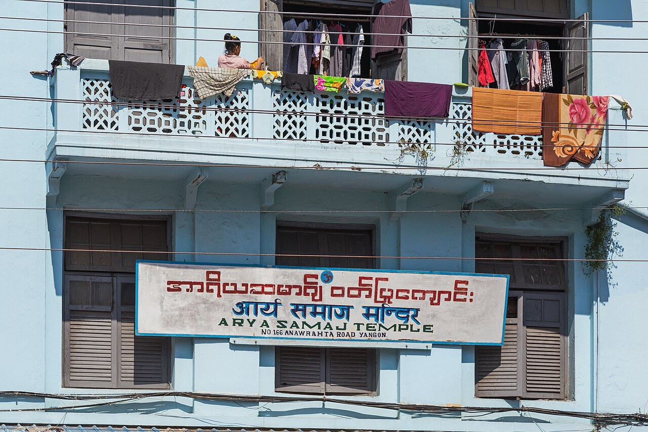 A pale-blue building in Yangon carries a trilingual signboard reading 'Arya Samaj Temple', with laundry strung above it.
