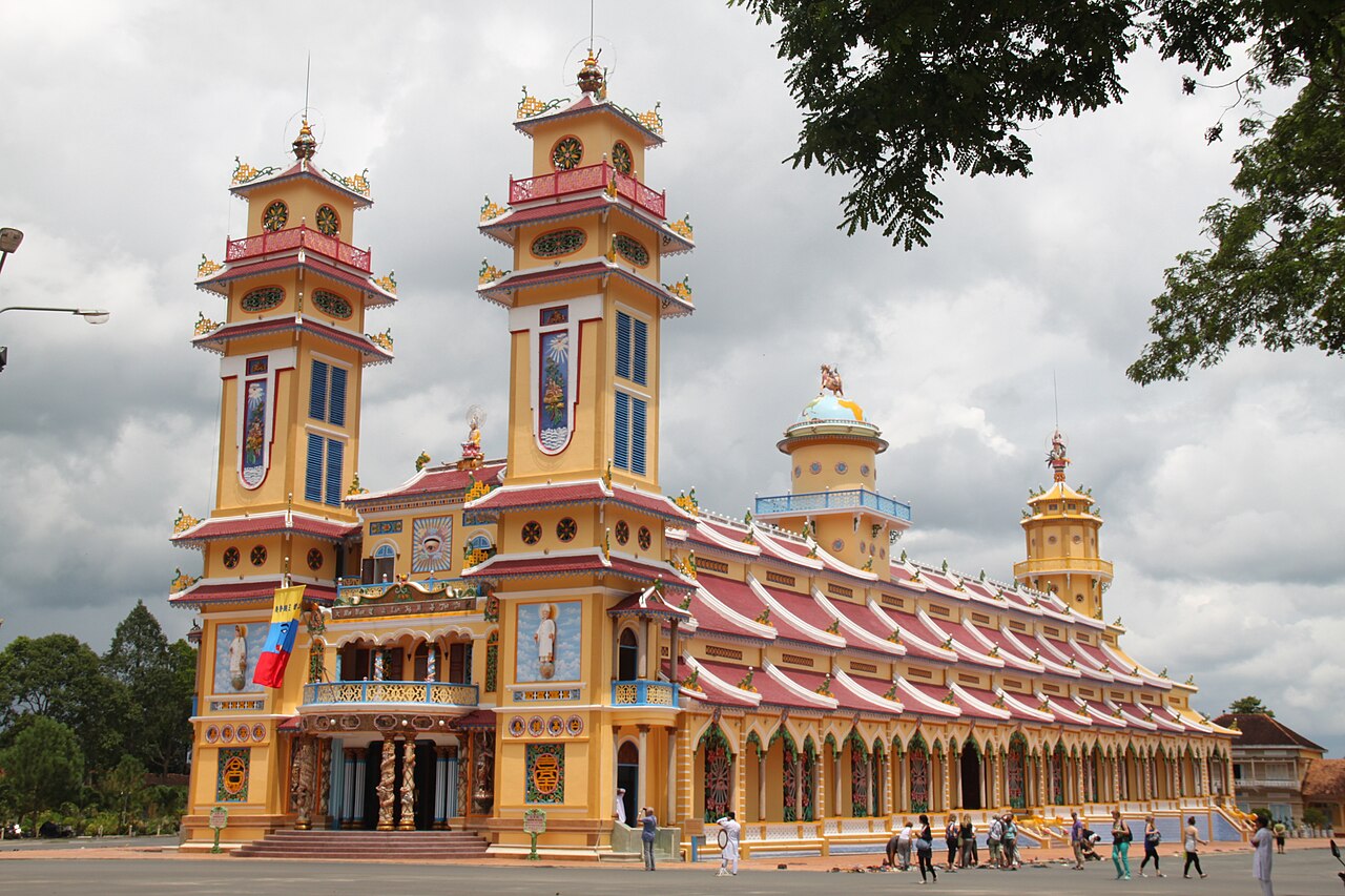 The Cao Dai Great Temple at the Holy See in Tây Ninh, Vietnam, its twin-towered, richly coloured façade under a cloudy sky.