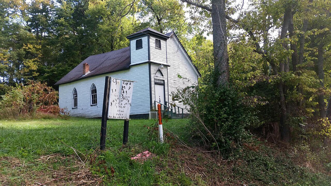 A small white CME church with a bell tower standing among trees in Winchester, Kentucky, its sign by the road.