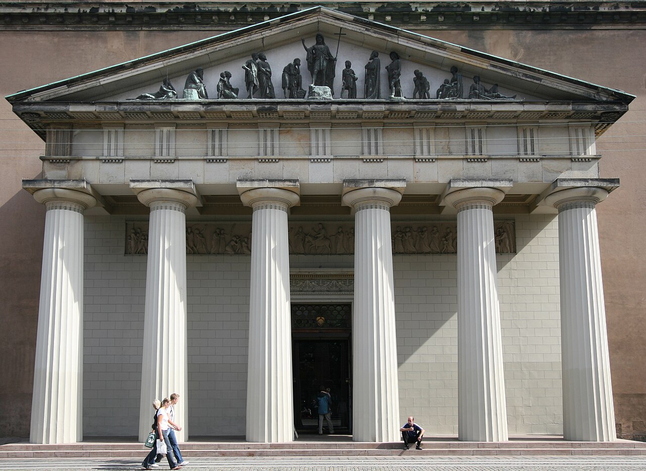 The columned neoclassical entrance of Copenhagen Cathedral (Vor Frue Kirke), two people walking past its steps.