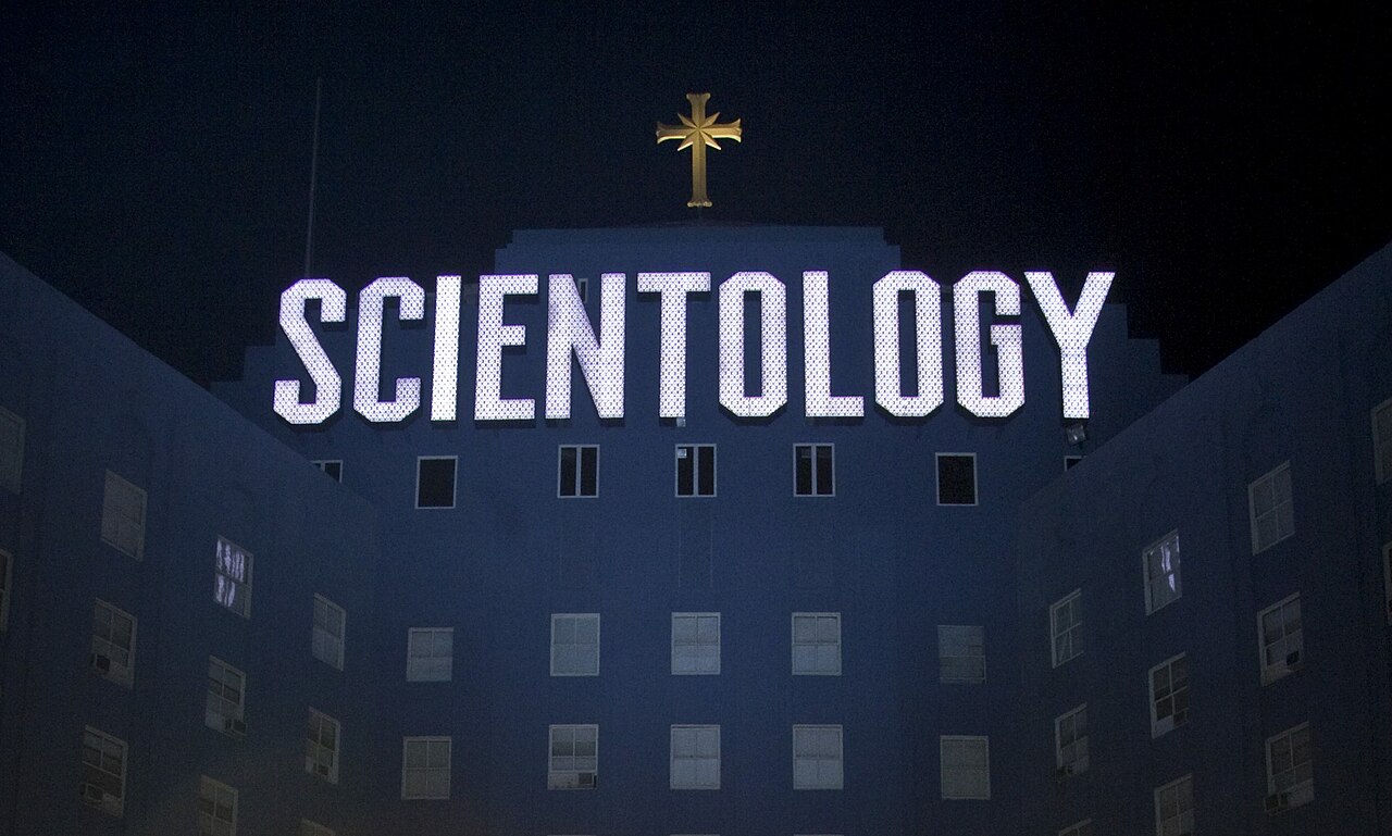 The Church of Scientology's Los Angeles building at night, its dark blue façade lit by a large SCIENTOLOGY sign and cross.