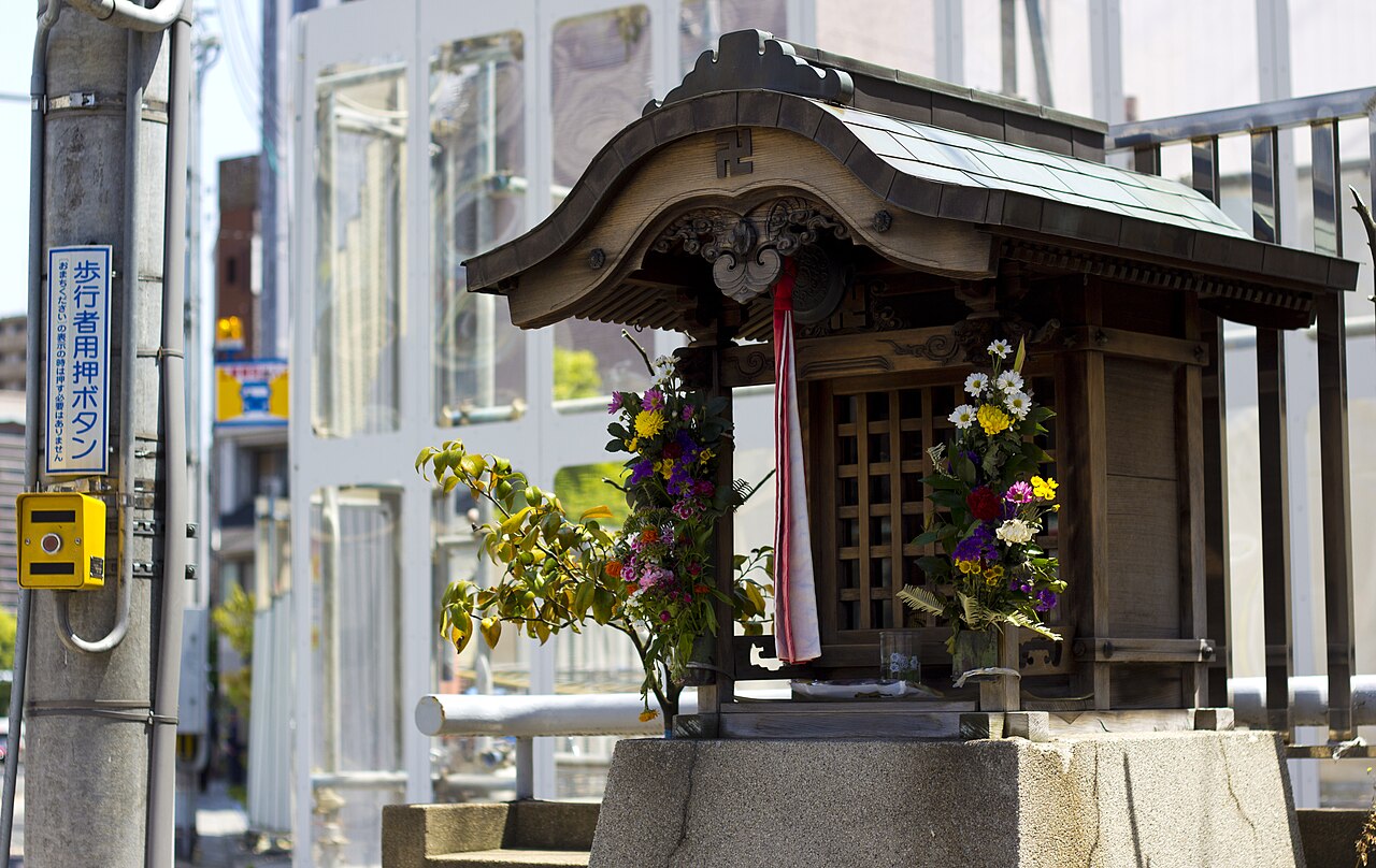A small wooden roadside hokora shrine in Kyoto, with flower offerings, standing beside a city street and a bus stop.