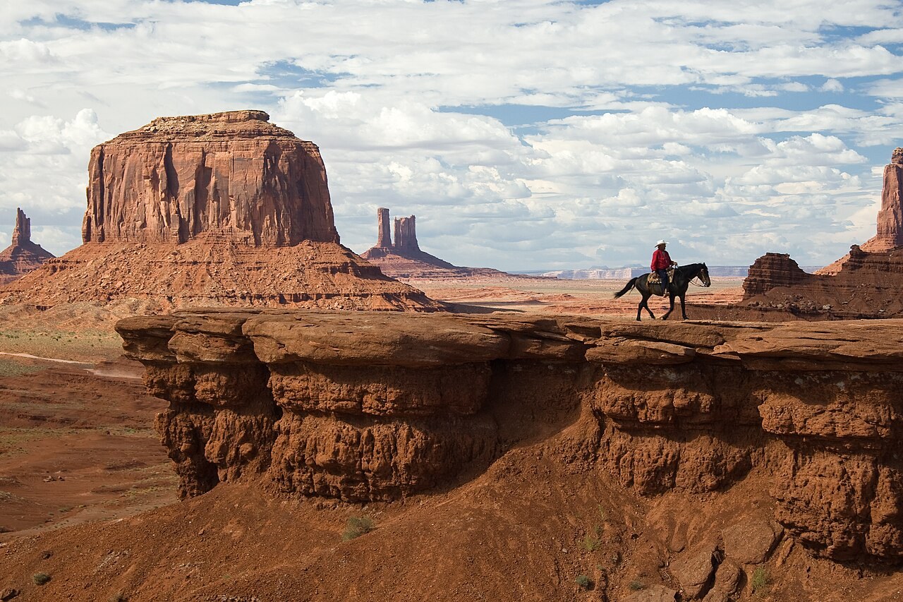 Red sandstone buttes at Monument Valley with a lone horse rider silhouetted on a rocky point.