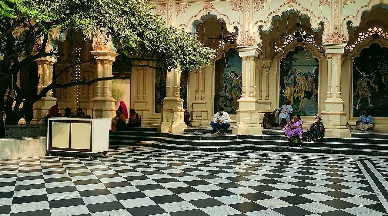 Inside the courtyard of ISKCON's Krishna Balaram Mandir in Vrindavan, arched walls painted with Krishna scenes surround a checkered marble floor.