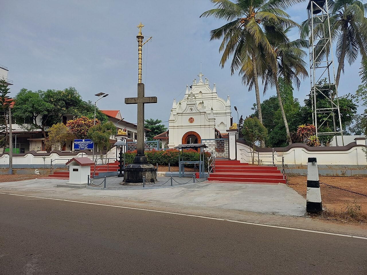 A white Malankara Orthodox church in Kerala behind its tall golden processional cross, palm trees around it.
