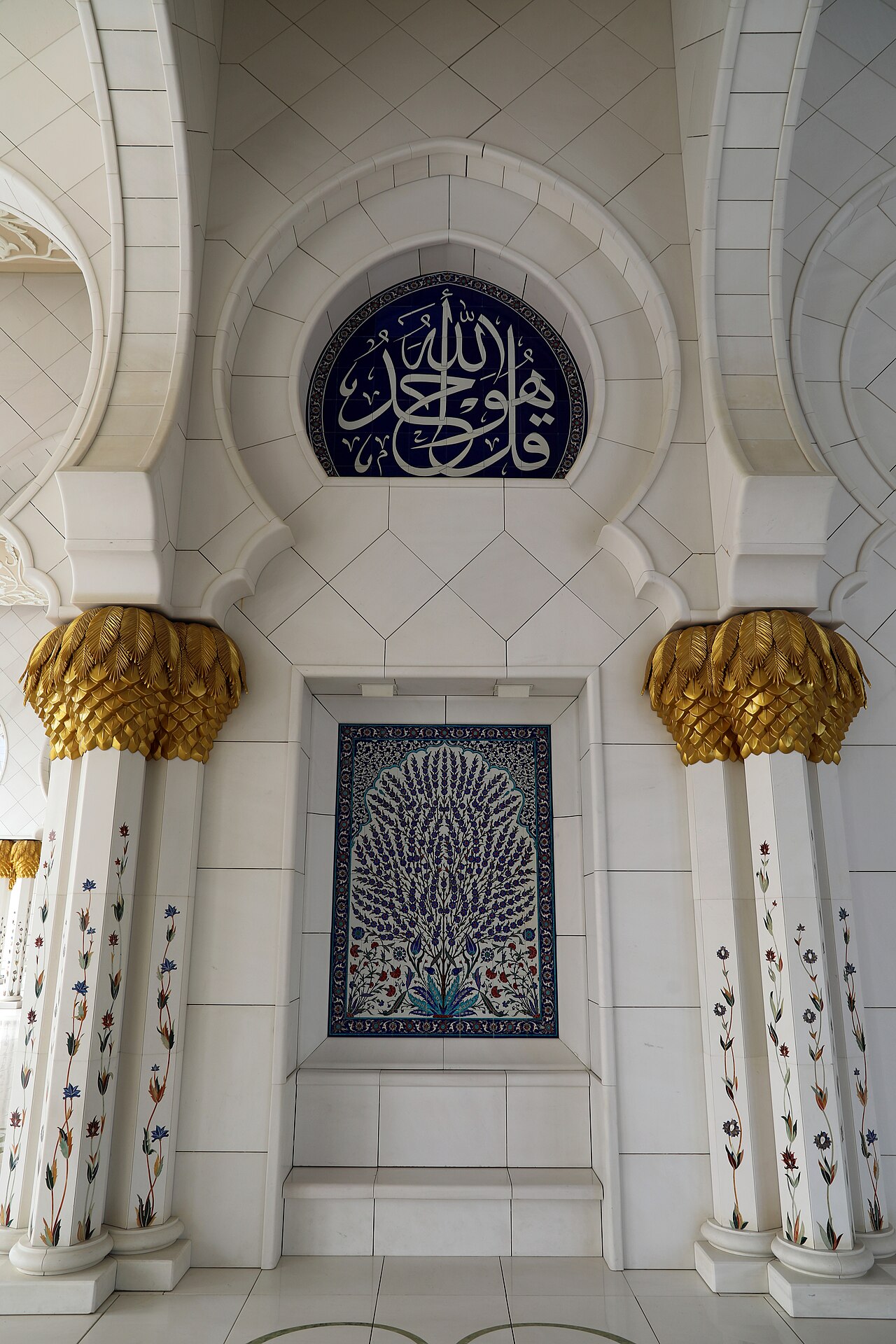 An arcade of the Sheikh Zayed Grand Mosque in Abu Dhabi: white marble columns with gilded capitals and floral inlay, a calligraphy roundel, and an Iznik-style tile panel.