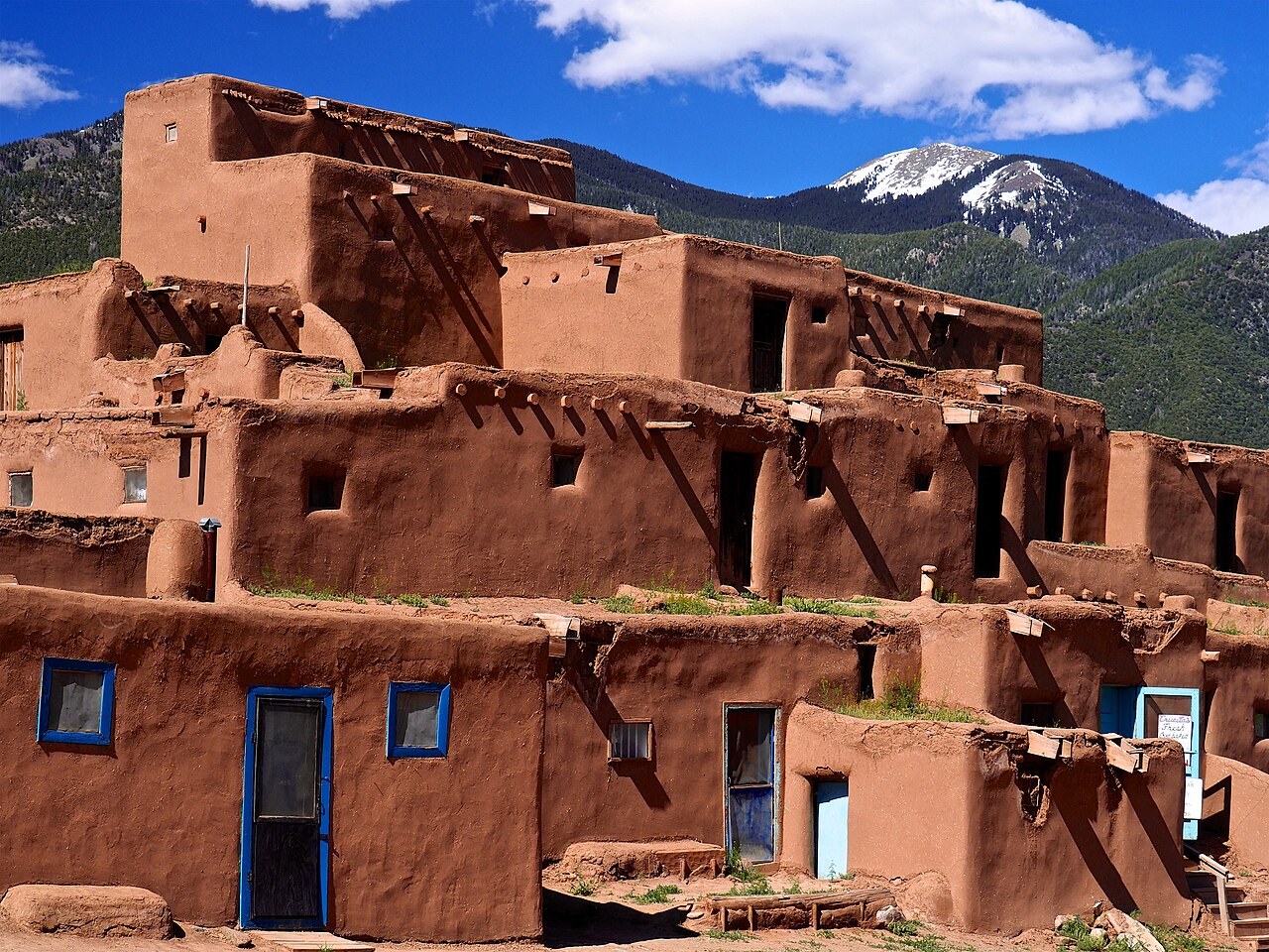 Terraced adobe houses with blue-trimmed doors at Taos Pueblo rise toward forested mountains.