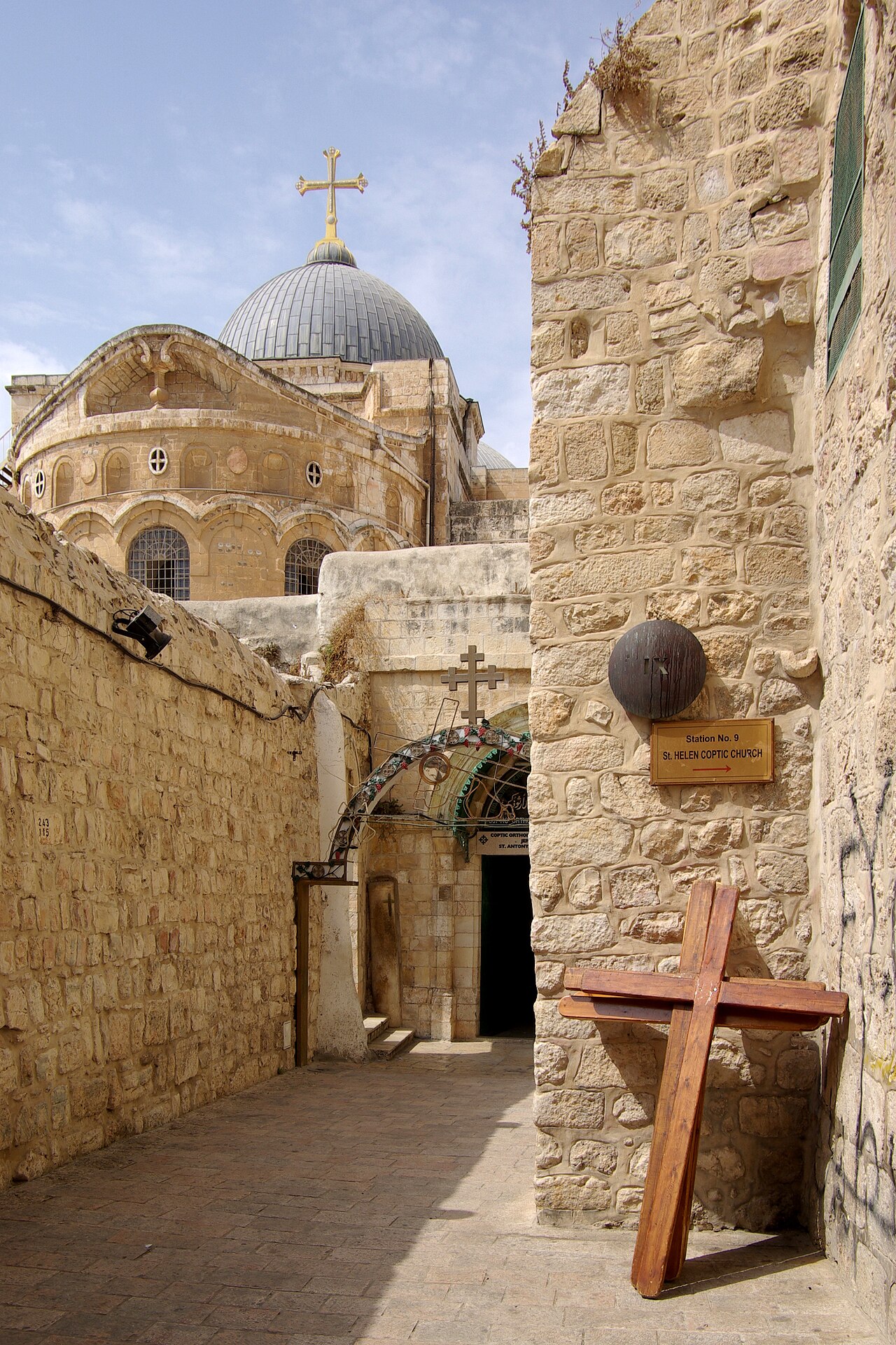 A stone alley beside the Church of the Holy Sepulchre in Jerusalem, a wooden cross in the foreground and the dome beyond.