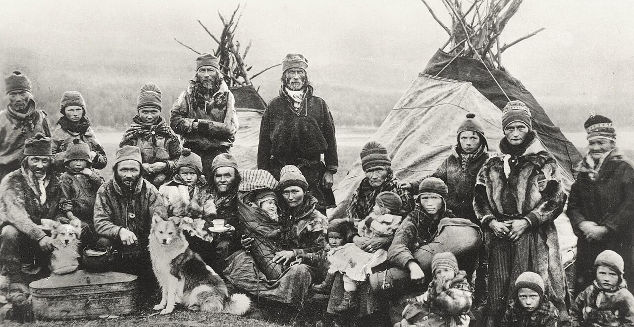 A black-and-white photograph from about 1900 to 1920 shows a Sámi group in traditional dress posed with dogs before lavvu tents.
