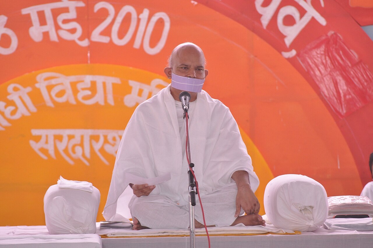 Acharya Mahashraman, head of the Terapanth order, sits in white robes with a Jain mouth-cloth before a microphone and an orange Hindi banner dated May 2010.