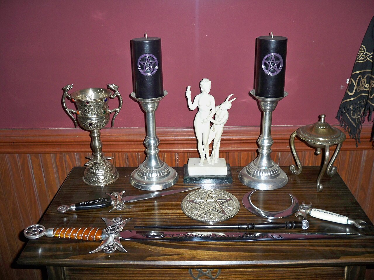 A Wiccan altar set with black pentacle candles, chalices, a small goddess figure, a pentacle disc, and ritual blades on a wooden table.