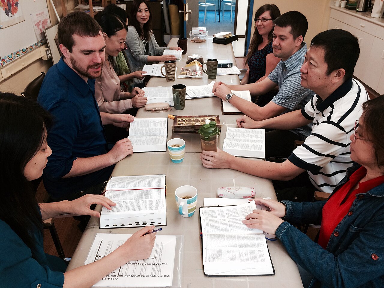 A Bible study group seated around a long table, open Bibles and study sheets in front of each person, in mid-discussion.