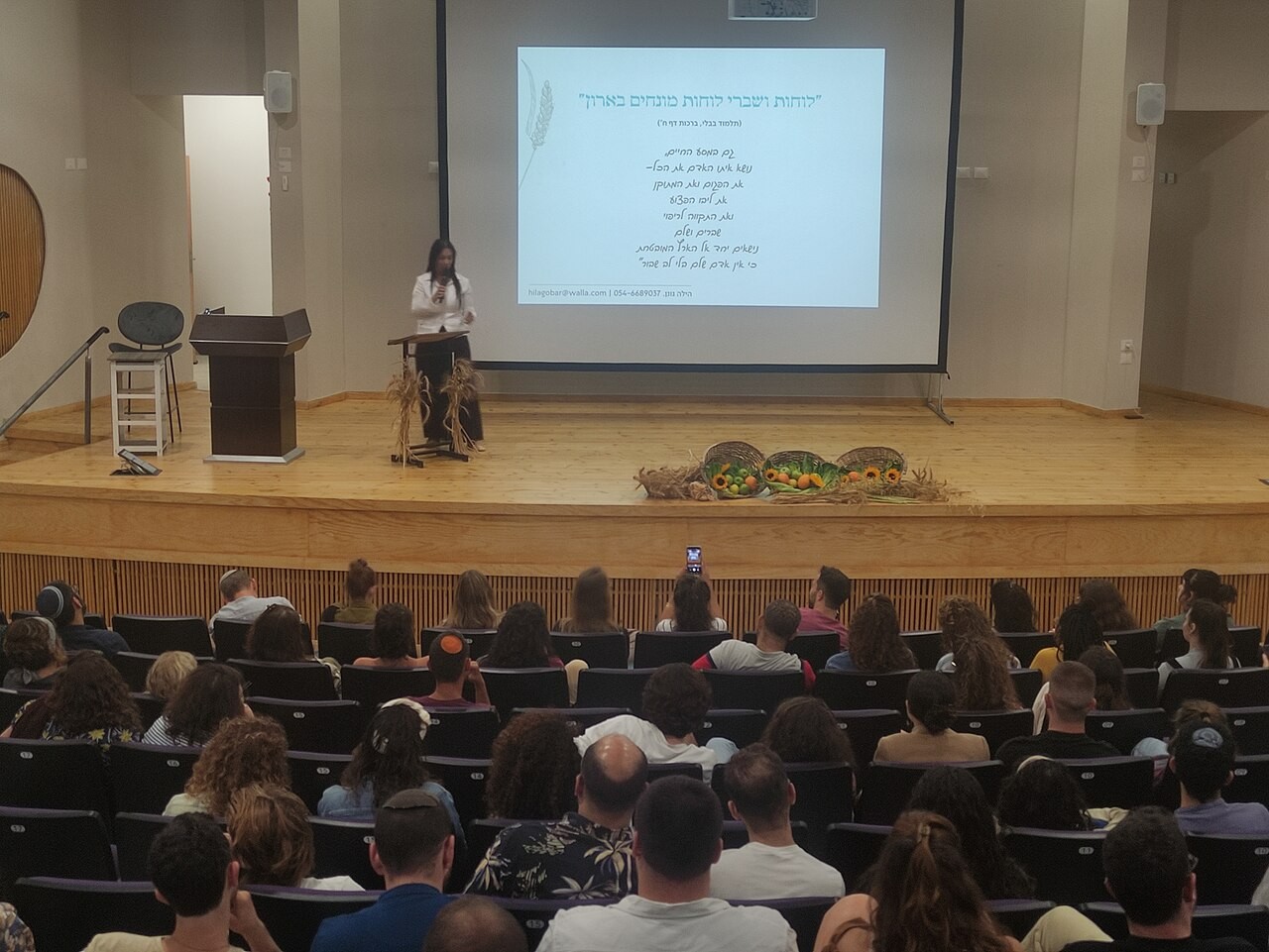 A modern Israeli beit midrash filled for a Shavuot study night, an audience facing a screen of Hebrew text while a teacher speaks from a lectern.