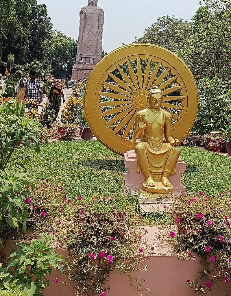A golden Buddha seated in teaching pose before a great golden wheel in the gardens at Sarnath.