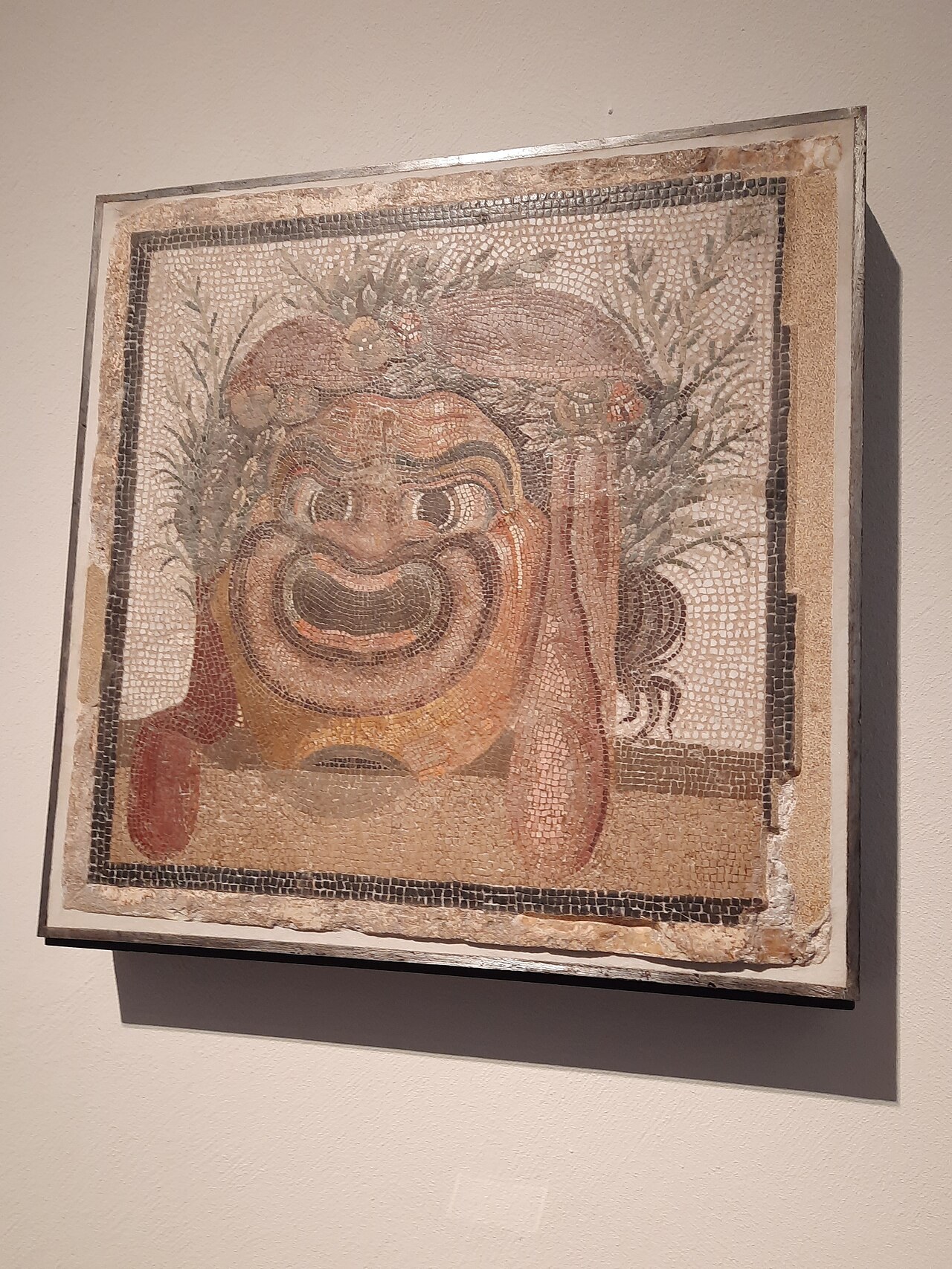 A Roman mosaic panel of a grinning theatre mask with an open mouth and garlanded hair.