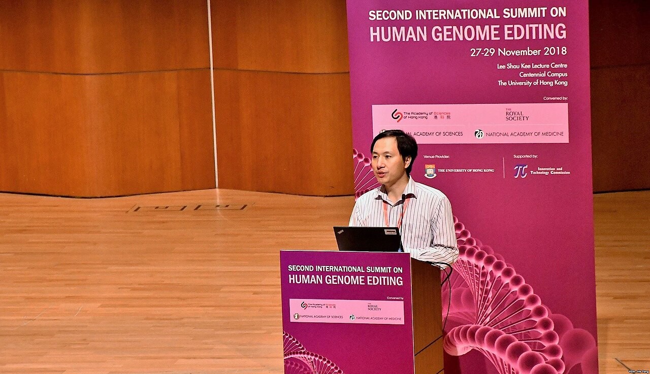 He Jiankui speaking at a podium before the banner of the 2018 Hong Kong summit on human genome editing.