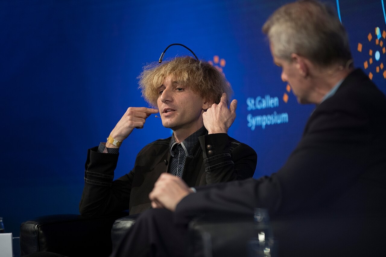 Neil Harbisson in conversation at the St. Gallen Symposium, his implanted antenna arching over his hair.