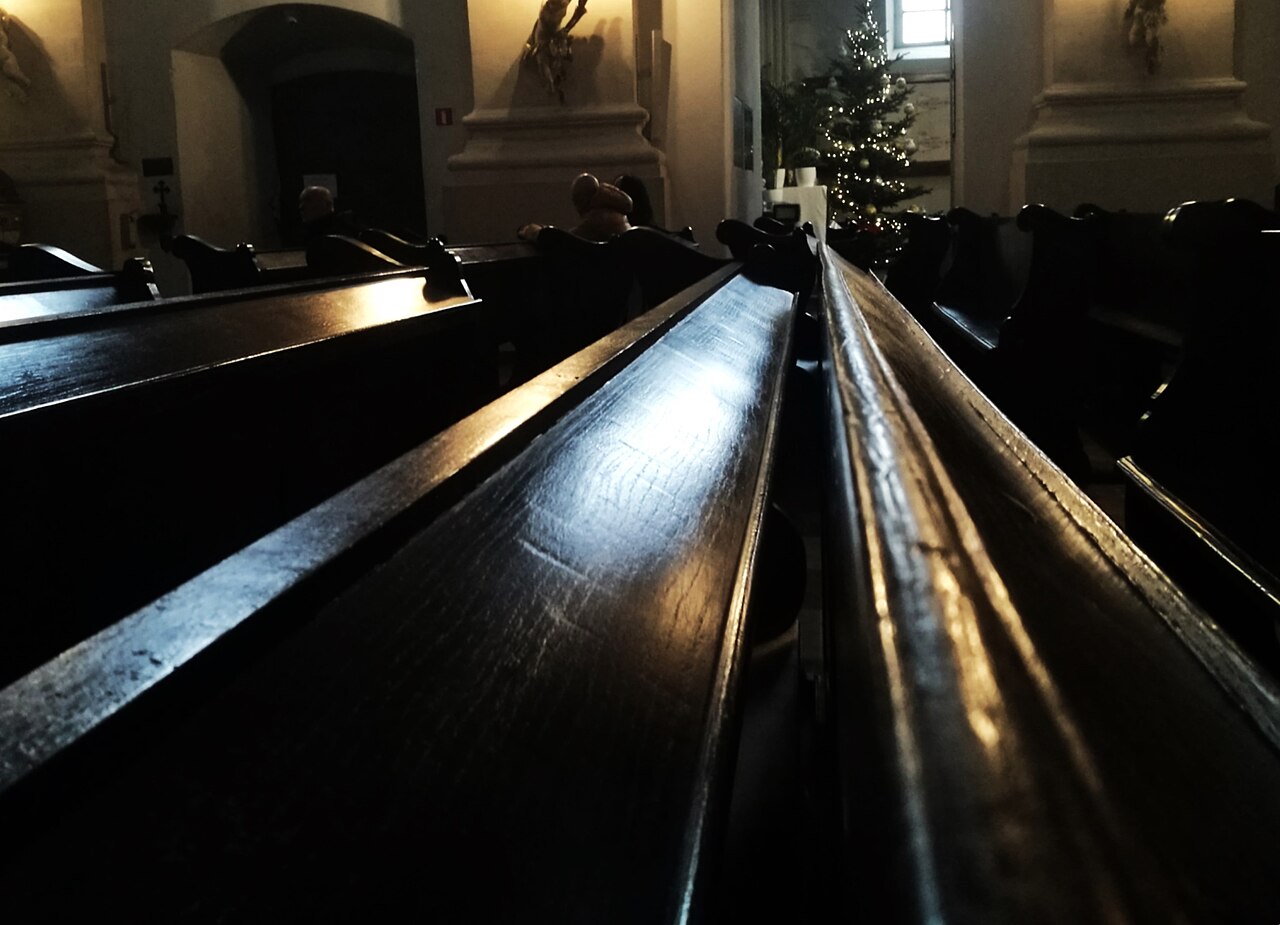 Long rows of dark, empty wooden pews recede through a dim church interior toward a lit Christmas tree.