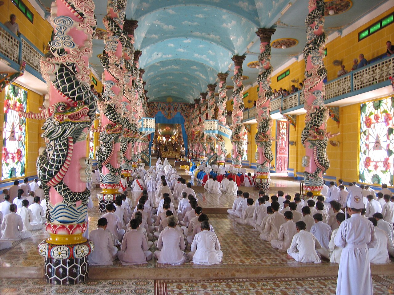 The interior of the Cao Dai Great Divine Temple at Tay Ninh during prayer: rows of white-robed worshippers between pink dragon-wrapped columns beneath a sky-blue ceiling.