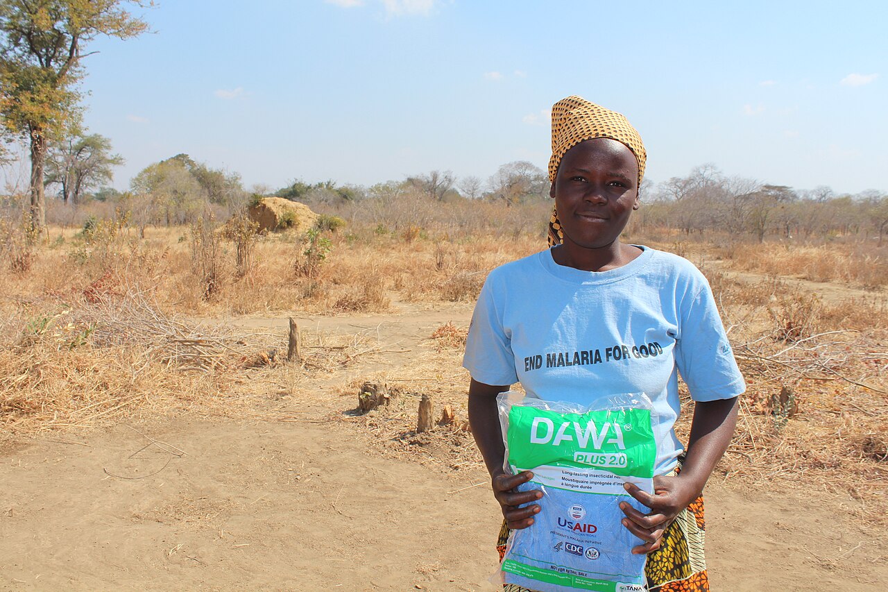 A woman in an END MALARIA t-shirt stands in dry bushland in Mbire, Zimbabwe, holding a still-packaged long-lasting insecticidal mosquito net from a distribution.