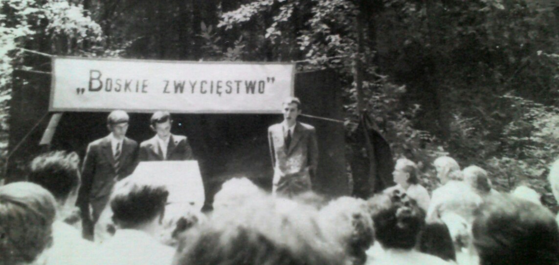 A black-and-white photograph of the 1973 'Divine Victory' assembly of Jehovah's Witnesses in Poland, speakers before a banner and a seated crowd.