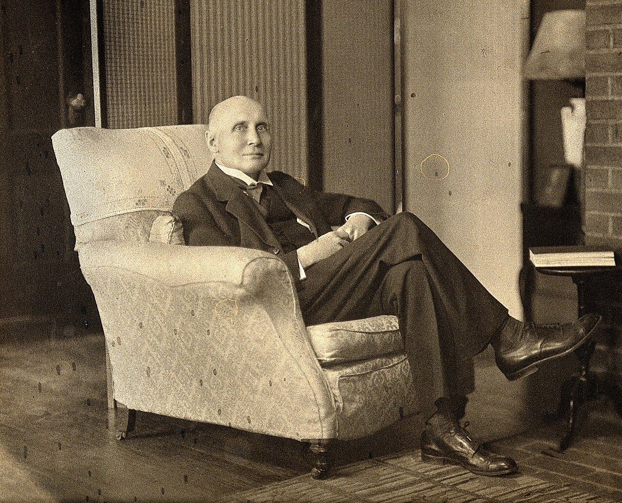 A sepia portrait photograph of the elderly Alfred North Whitehead in a three-piece suit, seated with legs crossed in a high-backed armchair.