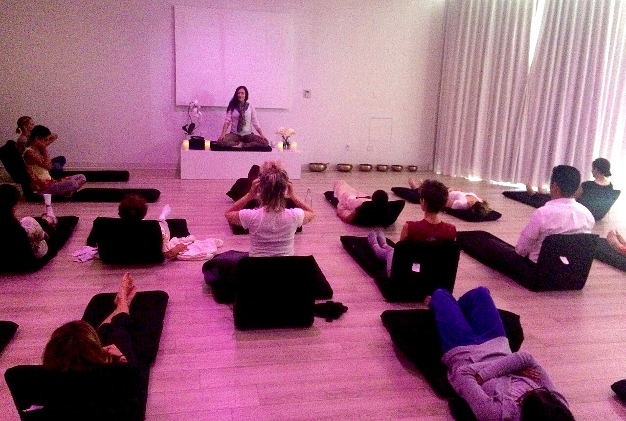 A guided meditation class in a bright, minimalist commercial studio: an instructor on a low platform, participants reclining on black floor cushions.