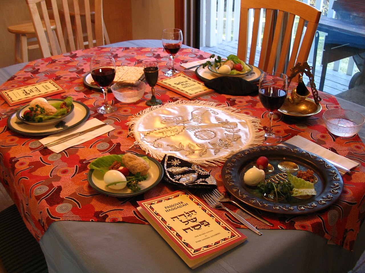 A home dining table set for a Passover seder: a seder plate with egg and herbs, matzah under an embroidered cover, glasses of red wine, and Haggadah booklets at each place.