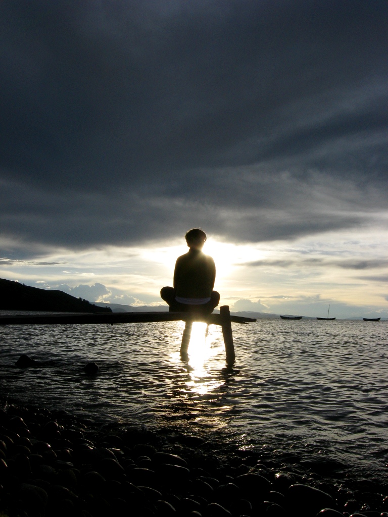 A lone person sits in lotus position on a wooden jetty over Lake Titicaca, silhouetted against the low sun on the water.