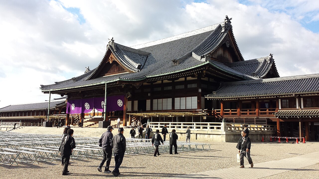 The Tenrikyo Church Headquarters Main Sanctuary in Tenri, a vast wooden hall with sweeping tiled roofs, seen across a gravel courtyard set with rows of folding chairs.