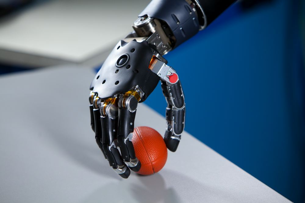 A black robotic prosthetic hand with articulated metal fingers gripping a small orange ball on a table.