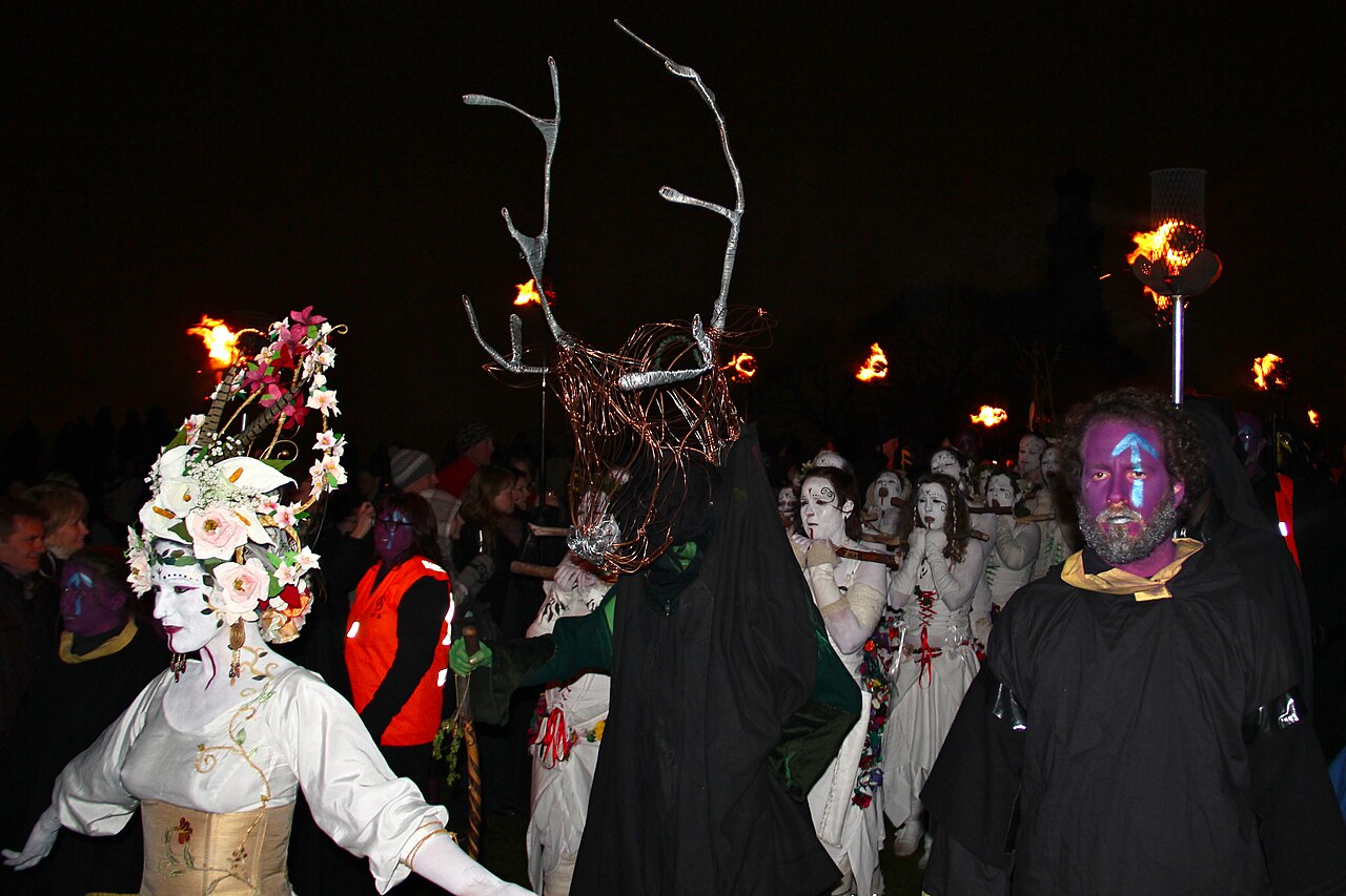 The nighttime procession of the Edinburgh Beltane Fire Festival: the flower-crowned May Queen, a figure in a towering antlered headdress, white-painted drummers, and torchbearers.